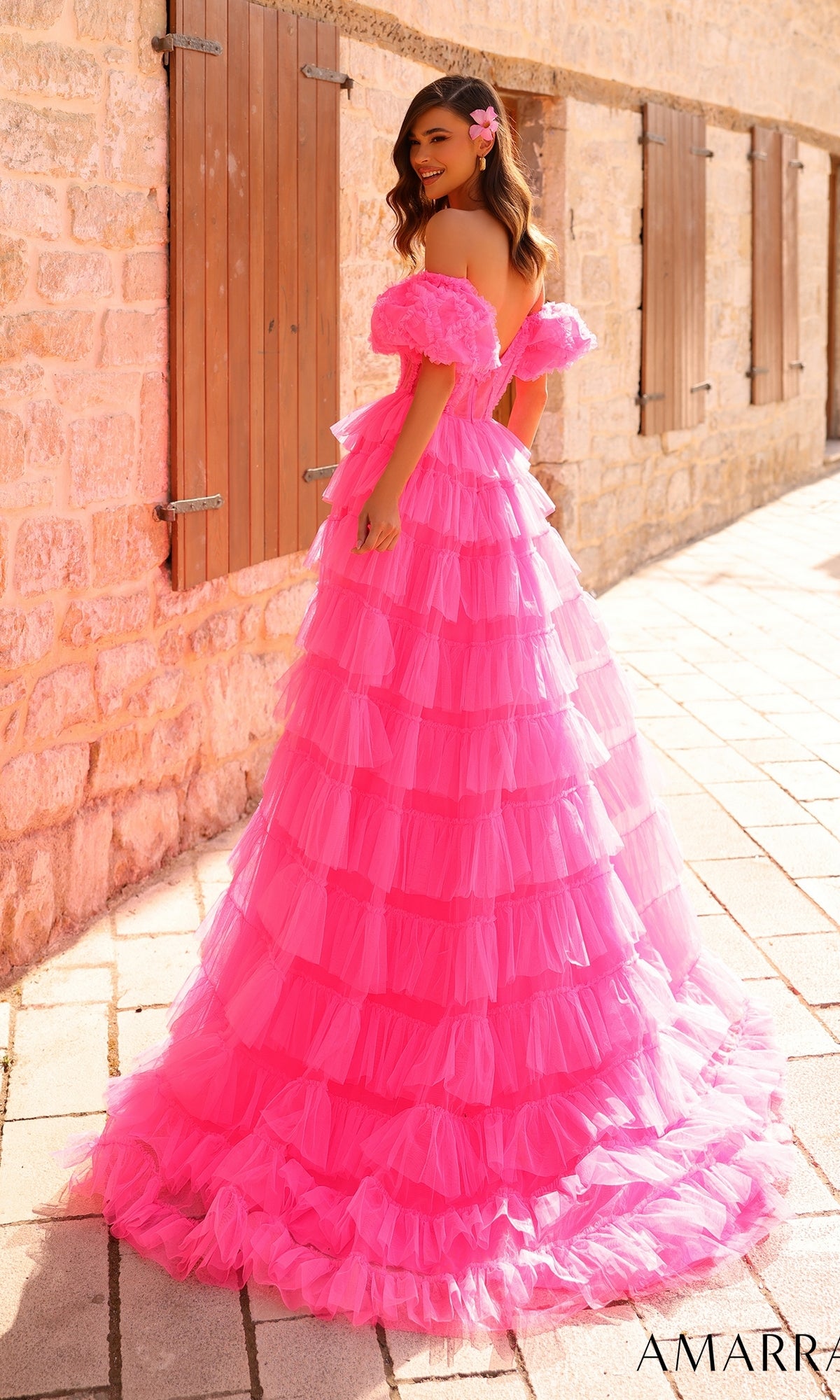 A woman stands outdoors before a stone wall, wearing the Amarra Long Prom Dress 94000—a bright pink ruffled tulle ball gown with off-the-shoulder puff sleeves and a flower in her hair.